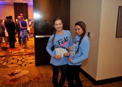 Two young women wearing blue shirts holding multiple event tickets, standing in a carpeted hallway with people in the background.