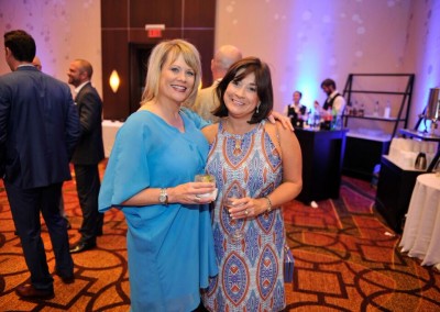 Two women smiling and holding drinks at a formal event with other guests in the background.