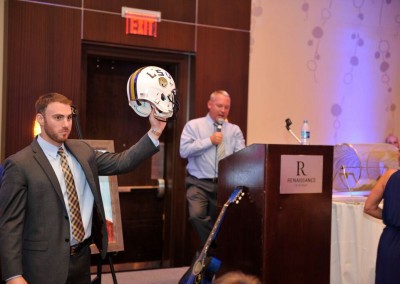 A man holding up a football helmet at an event while another man speaks into a microphone at a podium.