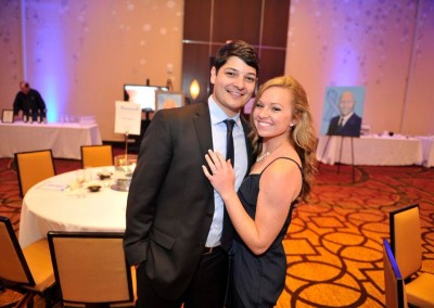 A couple smiling and embracing at a formal event in a room with round tables and a promotional banner in the background.