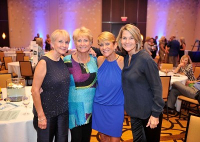 Four women posing together at a formal event, smiling in a banquet hall with tables and chairs in the background.