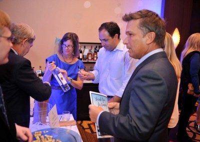 People interacting at a social event while a man pours a drink from a bottle into a glass at a booth labeled "taste of the universe.