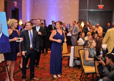 Networking event in a ballroom with people mingling and holding drinks, while a woman in a blue dress addresses a colleague.