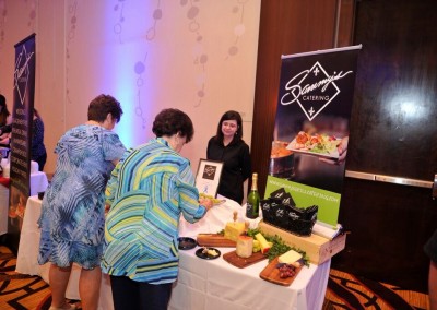Two women view a catering display table attended by a female vendor at a business event.