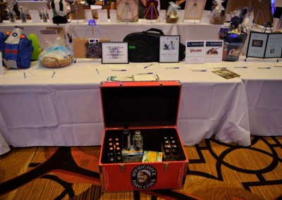 A red suitcase with liquor and snacks on display at a charity auction event, surrounded by other auction items on tables.