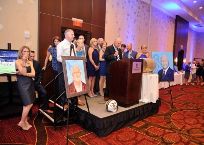 Group of people standing on a stage during a formal event, with a man speaking at a podium and portraits displayed nearby.