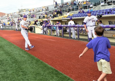 A young boy runs excitedly towards baseball players awaiting in the dugout during a pre-game warm-up on a synthetic turf field.