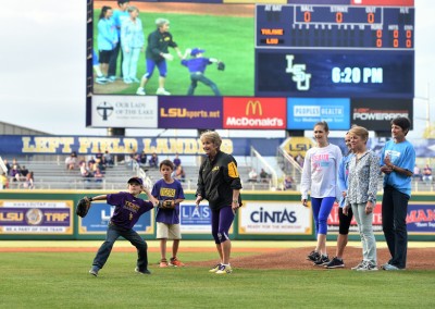 A boy throws the first pitch at an lsu baseball game with spectators and scoreboard reading "0-0" in the background.