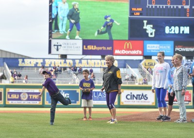 Young boy in mid-throw on a baseball field with onlookers, including an elderly woman, during a pre-game event.