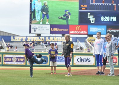 A child throws the ceremonial first pitch at a baseball game, with spectators and scoreboard in the background.