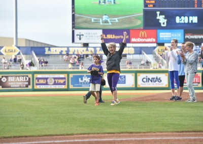 A woman and a young boy running on a baseball field, smiling and waving, with spectators in the background.