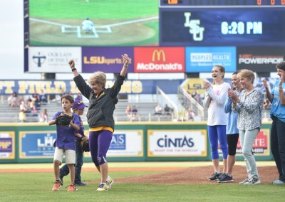 A woman in a purple lsu sweatshirt cheers on a baseball field with a boy next to her, as spectators applaud from behind.