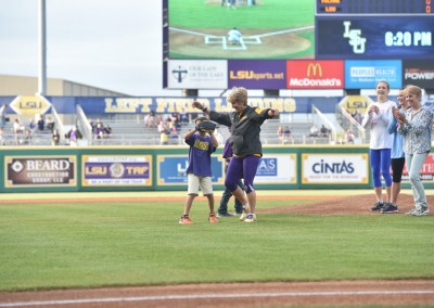 A young boy in a purple lsu shirt dances with an adult on a baseball field as spectators watch and cheer.