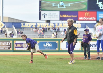 A young boy throws a baseball on a field while a woman in lsu apparel watches, with other spectators in the background.