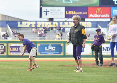 A young boy in a purple lsu shirt throws a baseball on a field, watched by an adult in lsu attire and other spectators.