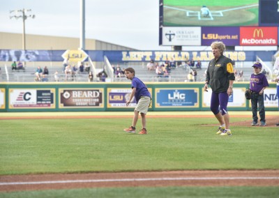 A child throws a baseball on a field at an event while spectators and a coach watch in the background.