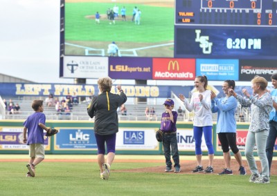 Young boy in baseball gear runs to his family on a field, with spectators and a large scoreboard in the background at an lsu baseball game.