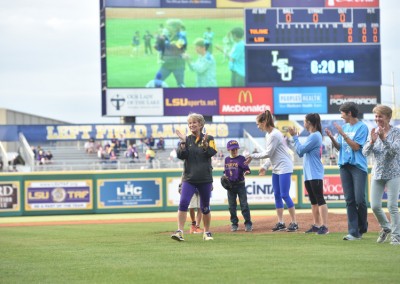 A young child is being honored on a baseball field while spectators and a mascot applaud; a scoreboard is visible in the background.