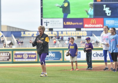 A woman in lsu apparel clapping on a baseball field, with several people and a child nearby, and a large digital scoreboard in the background.