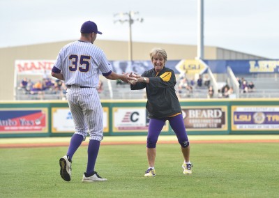 An older woman in a yellow and purple jacket high-fives a younger baseball player in a striped purple and white uniform on a baseball field.
