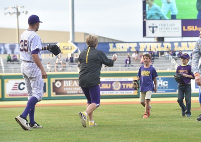 An older woman runs playfully on a baseball field towards a player as a young boy watches, all surrounded by stadium ads and spectators.