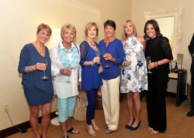 Six mature women smiling and holding wine glasses at an indoor gathering.