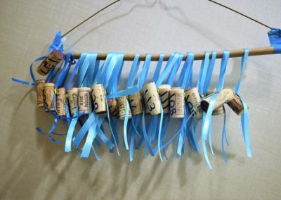 A handmade wind chime made from wine corks, blue ribbons, and a stick, suspended against a pale background.