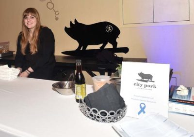 Woman at a promotional booth for city pork, featuring culinary supplies, a basket of bread, and a beverage, with a black pig silhouette on the wall.