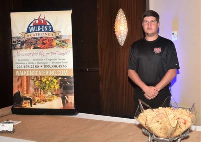 Man standing beside a catering banner and a table with a dish at a walk-on's catering event.