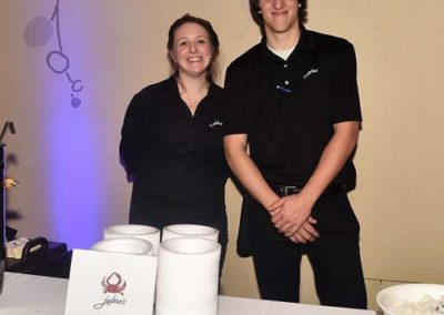 Two caterers in black uniforms standing behind a table with food containers at an event.