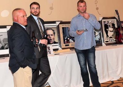 Three men standing next to a display with memorabilia, one holding a wine glass, another a smartphone, in a banquet hall.