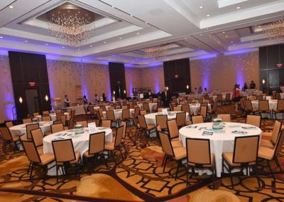 Interior of a banquet hall with round tables, chairs, and chandeliers, set up for an event.