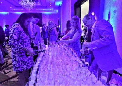 People serving champagne at a formal event with a table lined with glasses under blue lighting.