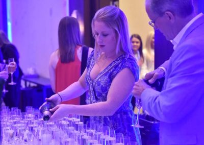 A woman pouring champagne into glasses at an event with attendees in the background.