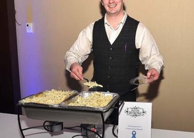A smiling man serving pasta at a catering event, standing behind chafing dishes on a table with an event sign.
