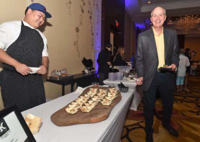 A chef and a guest smiling at a food tasting event, with a wooden board of appetizers displayed on a table.