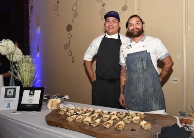 Two chefs standing behind a food sampling table at an event, smiling, with a display of appetizers in front of them.