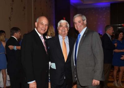 Three smiling men in suits posing together at a formal event with a blue-lit background.