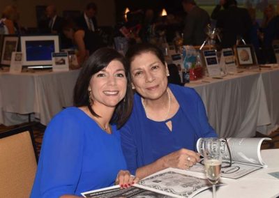 Two women in blue dresses smiling at a table during an event, with newspapers and a champagne glass in front of them.