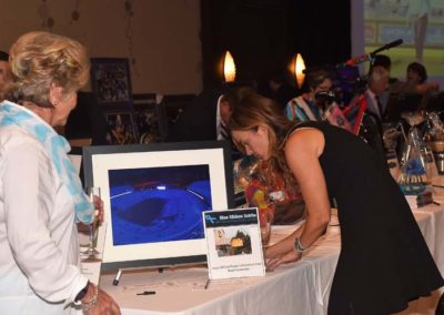 Two women examining a silent auction item at a formal event, with other attendees and auction items in the background.