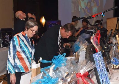 Two people examining items at a charity auction event, with others in the background and various gift baskets on display.