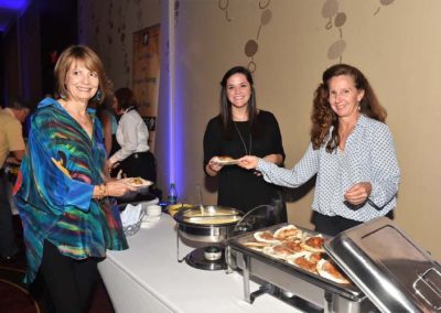 Three women smiling while serving themselves food at a buffet in a banquet hall.