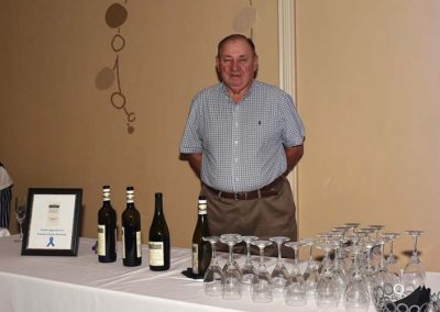 Man standing behind a table with wine bottles and glasses, smiling in a room with awards on the table.