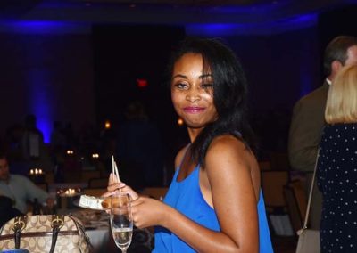 A woman in a blue dress holding a champagne glass at a formal event, smiling at the camera with people in the background.