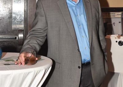 Man in a gray suit and blue shirt holding a drink, standing by a table at a formal event.
