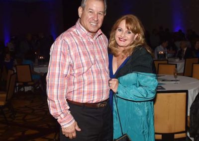 Two adults smiling at a formal event, one in a pink checkered shirt and the other in a teal dress.