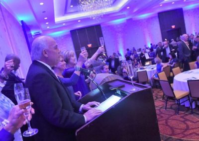 Man at podium giving a toast at a formal event with attendees raising glasses in a banquet hall with blue lighting.