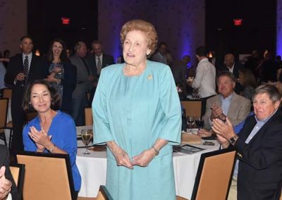 An elderly woman in a teal dress stands smiling at a formal event, while seated guests around her applaud.