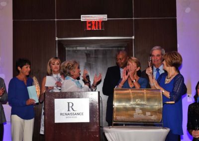 Group of people applauding a woman speaking at a podium during an event in a hotel ballroom.