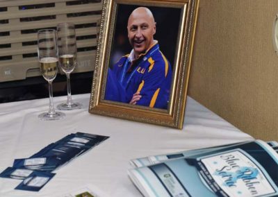 A framed portrait of a smiling man in a blue and yellow uniform on a table with champagne glasses and brochures.